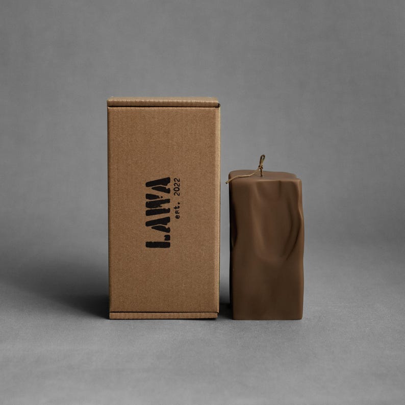 May include: A brown square candle with a textured surface and a wick, next to a cardboard box. The box has the word "LAWA" printed on it, with "est. 2022" below it.