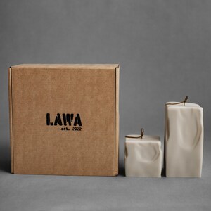 May include: A square cardboard box with the text "LAWA est. 2022" in black. Two off-white, textured candles of different sizes are next to the box. The candles have a square shape and are tied with a brown string.