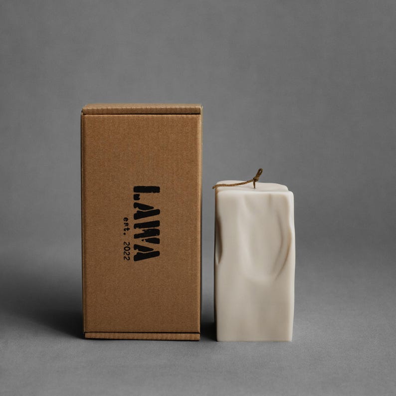 May include: A cream-colored, square candle with a textured surface and a brown wick, next to a brown cardboard box. The box has the word "LAWA" and "est. 2022" printed in black. The background is a solid gray.