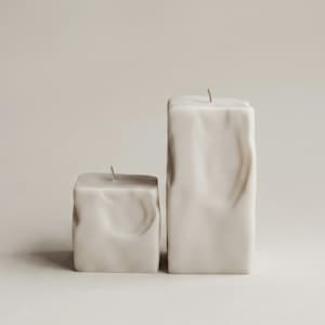 May include: Two off-white, square-shaped candles with textured surfaces. One candle is shorter and wider than the other. Each candle has a single, thin wick at the top, suggesting they are ready to be lit. The candles are set against a neutral background.