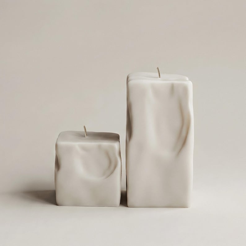 May include: Two off-white, square-shaped candles with textured surfaces. One candle is shorter and wider than the other. Each candle has a single, thin wick at the top, suggesting they are ready to be lit. The candles are set against a neutral background.