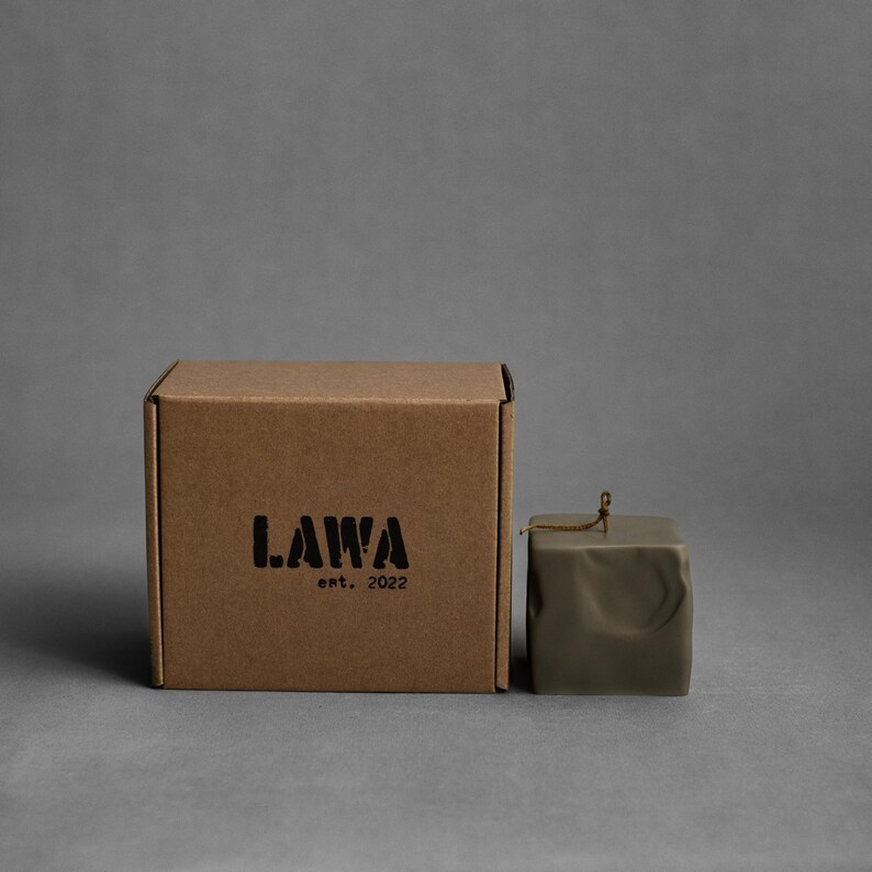May include: A square, olive-green candle with a textured surface and a small, decorative string. Beside it is a brown cardboard box with the black text "LAWA est. 2022". The background is a solid gray.