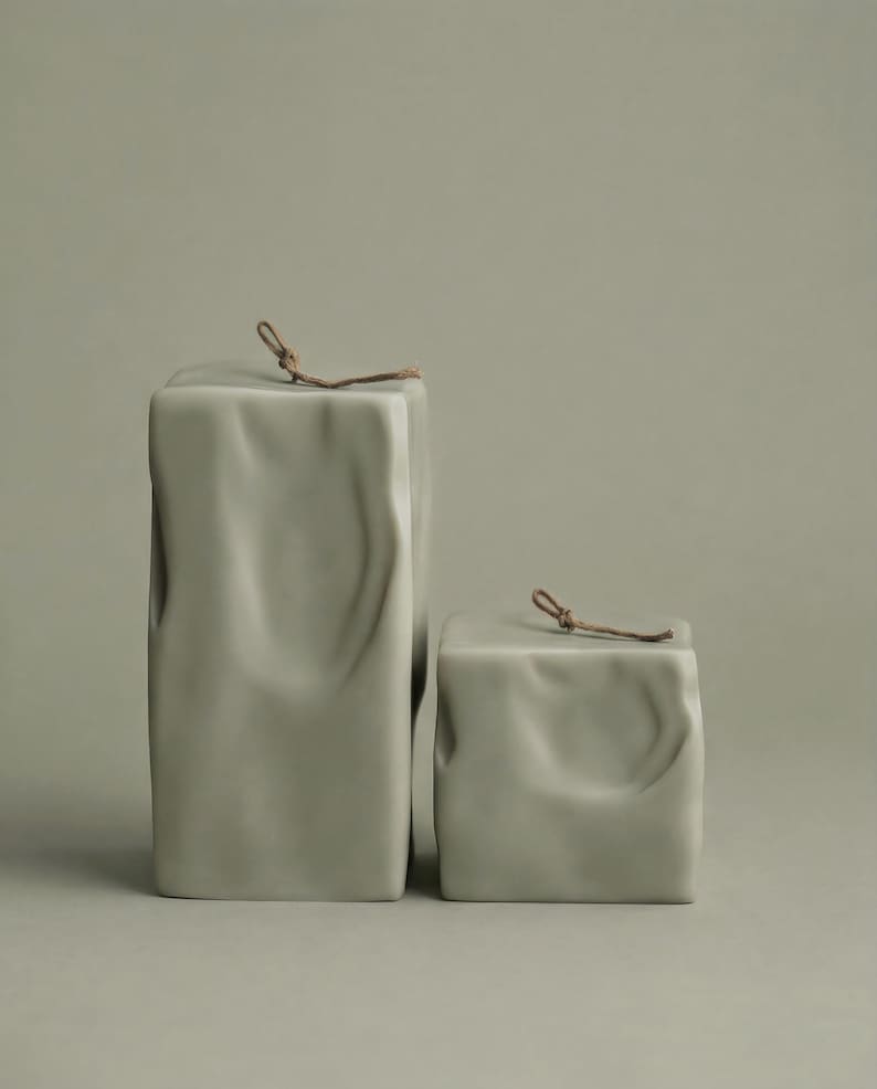 May include: Two light green, rectangular ceramic containers with a textured, draped appearance. The larger container is taller than the smaller one. Each has a small, brown, knotted cord attached at the top. The background is a solid, muted green.
