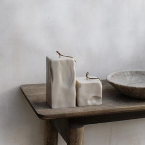 May include: Two off-white, textured block candles with a rustic aesthetic. Each candle has a small, brown wick. The candles are displayed on a wooden table, alongside a stone bowl. The text "RAW BLOCK | SET OF 2" is visible.