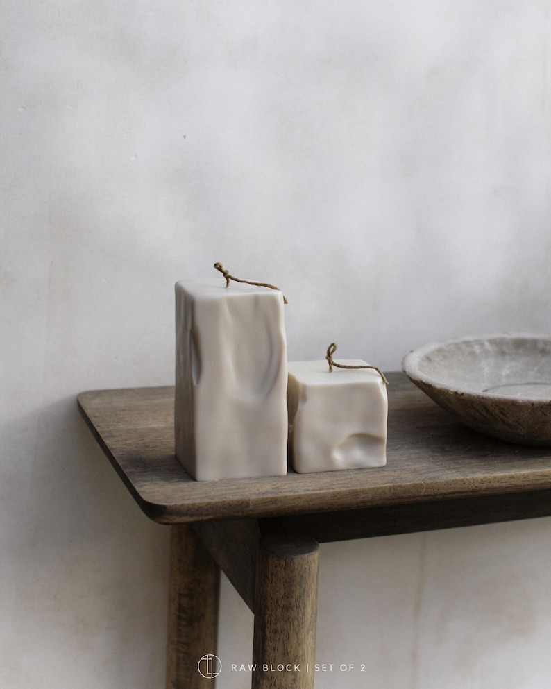 May include: Two off-white, textured block candles with a rustic aesthetic. Each candle has a small, brown wick. The candles are displayed on a wooden table, alongside a stone bowl. The text "RAW BLOCK | SET OF 2" is visible.