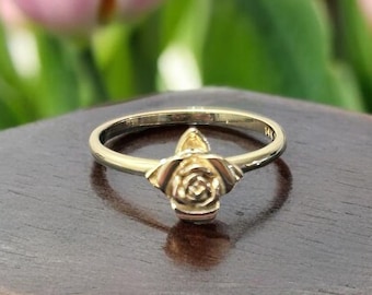 Può includere: Un anello d'oro con un design a forma di rosa. L'anello è su una superficie di legno marrone scuro.