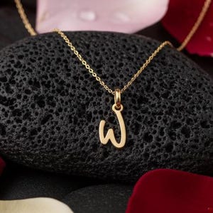 Könnte beinhalten: Eine goldfarbene Halskette mit einem Anhänger in Form des Buchstabens „W“ liegt auf einem schwarzen, porösen Stein. Rote, rosa und weiße Rosenblätter umgeben den Stein, vor einem Hintergrund aus schwarzen Steinen.