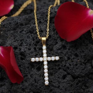 May include: A gold-colored necklace with a cross pendant encrusted with clear gemstones. The cross is centered on a dark, textured surface, surrounded by red rose petals. The necklace chain is delicate.