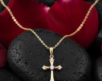 May include: A gold-colored cross pendant necklace with small, clear gemstones. The necklace is displayed on a black stone surface, surrounded by red rose petals.