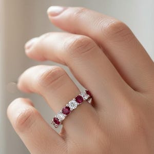 May include: A close-up of a ring featuring alternating round-cut rubies and clear gemstones. The ring is set in a silver band and is worn on a person's finger. The gemstones are vibrant red and clear.