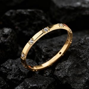 May include: A gold-colored ring with small, clear gemstones set along the band. The ring is displayed on a bed of dark, textured rocks. The ring's design is simple and elegant, with a polished finish.