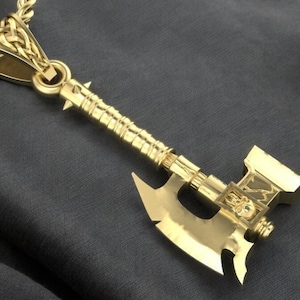 May include: A gold-colored axe pendant with a detailed design. The axe has a skull-shaped detail on the head and a braided rope chain.