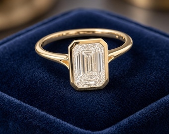 Könnte beinhalten: Ein Goldring mit einem rechteckigen Smaragdschliff-Edelstein in einer glatten Zargenfassung. Die Ringschiene besteht aus poliertem Gelbgold mit einem schlanken Profil, das sich zum Stein hin verjüngt und ein klares Design ergibt.
