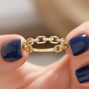 May include: A gold chain-link ring with small diamonds set in the center links. The ring is held between two hands with dark blue nail polish. The ring is marked with "14K".