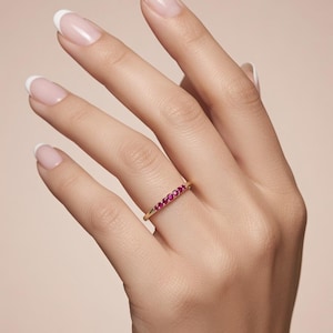 May include: A thin gold band ring featuring a row of five small, round, vibrant pinkish-red gemstones set horizontally across the top. The ring has a polished metallic finish and a simple, slender design that sits comfortably on the finger.