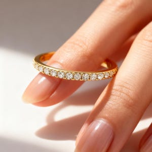 May include: A gold ring with a band of small, clear gemstones. The ring is held between two fingers, showcasing the ring's design and the light reflecting off the gemstones. The ring appears to be a wedding band or an eternity ring.