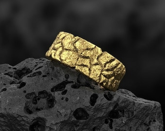 May include: A gold-coloured ring with a textured surface, resting on a dark grey, porous rock. The ring's design features a pattern of irregular shapes, giving it a unique, organic appearance. The background is a blurred black.