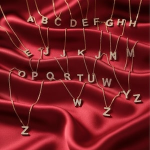 Puede incluir: Collares dorados con colgantes de iniciales, cada letra incrustada con pequeñas piedras brillantes. Los collares se exhiben sobre una tela de satén rojo intenso, creando una presentación lujosa y elegante. Se ven las letras de la A a la Z.