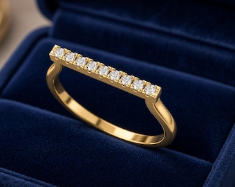 May include: A gold ring featuring a horizontal bar set with nine small, round, clear stones. The ring is displayed inside a dark blue velvet jewelry box. The band is smooth and polished, connecting to the ends of the straight bar setting.