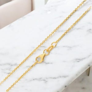 May include: A gold-coloured chain necklace with a lobster clasp. The chain is made of small, round links. The necklace is displayed on a white and grey marble surface.