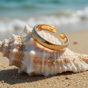 Könnte beinhalten: Ein goldfarbener, verstellbarer Ring, markiert mit "14K", liegt auf einer Muschel an einem Sandstrand. Der Ring ist offen, was eine Größenanpassung ermöglicht. Der Hintergrund zeigt das Meer und einen sonnigen Tag.