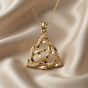 Pode incluir: Um pingente de tom dourado com um design de nó triquetra entrelaçado, suspenso por uma delicada corrente de tom dourado a condizer. O pingente tem uma superfície polida e reflexiva com linhas curvas suaves que formam uma forma triangular.