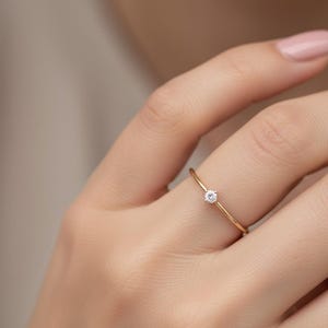 Może przedstawiać: Delikatny złoty pierścionek z małym, okrągłym, przezroczystym kamieniem szlachetnym pośrodku. Pierścionek jest na palcu, prezentując jego prosty i elegancki design. Opaska jest cienka i wypolerowana, odbijając światło.