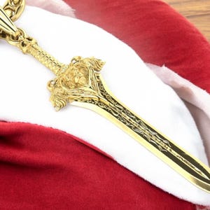 May include: A gold-coloured sword pendant with a lion head design. The pendant is attached to a thick gold chain.