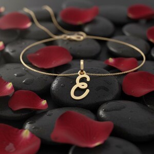 May include: A gold-colored necklace with a pendant shaped like the letter "E" rests on a black stone. Red rose petals are scattered around the necklace and stones, creating a romantic and elegant display.