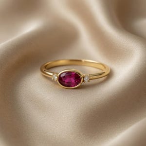 May include: A gold ring featuring a large oval ruby gemstone and two small diamond accents. The ring is set on a slender gold band. The ring is displayed on a satin fabric.