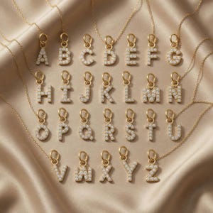 Könnte beinhalten: Eine Sammlung goldfarbener Anhänger-Halsketten mit Initialen, die jeweils einen anderen Buchstaben des Alphabets zeigen. Die Buchstaben sind mit kleinen, funkelnden Steinen besetzt. Die Halsketten liegen auf einer beigen, satinartigen Oberfläche.