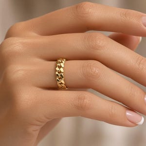 May include: A gold ring featuring a continuous curb chain link design. The band is crafted with polished, interlocking metal links that create a textured, repeating pattern around the entire circumference of the circular jewelry piece.
