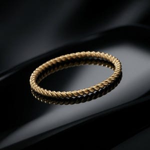 Puede incluir: Un anillo fino y circular hecho de metal dorado, que presenta una textura de cuerda trenzada continua en toda la banda. El anillo tiene un acabado pulido que capta la luz, creando un patrón en espiral distintivo a lo largo de su circunferencia.