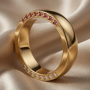 May include: A gold ring with a polished finish, set with small red and clear gemstones. The ring is displayed on a cream-colored, silky fabric, creating a luxurious aesthetic. The gemstones are evenly spaced around the ring.