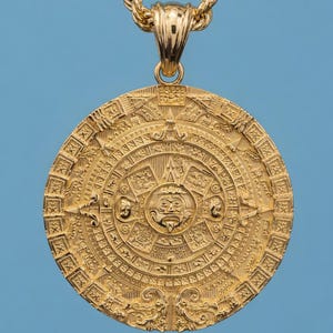 May include: A gold-colored pendant necklace featuring a detailed Aztec calendar design. The circular pendant is intricately carved with various symbols and figures. The necklace has a rope chain and a bail for the pendant.
