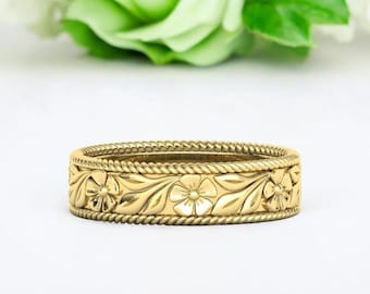 May include: A gold band ring with a floral design. The band is decorated with a rope-like pattern and features a row of stylized flowers with leaves.