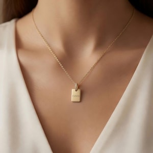 Puede incluir: Un collar de tono dorado con una cadena fina y un pequeño colgante rectangular. El colgante tiene una superficie lisa y pulida con el nombre "AURORA" grabado en una fuente sin remates. Una pequeña piedra clara está engastada en la argolla superior.