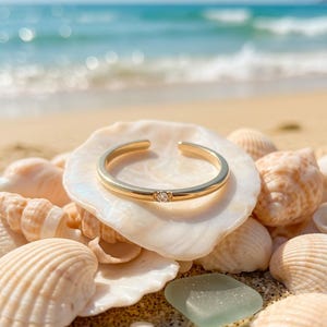 Peut inclure: Une bague ouverte dorée ornée d'une petite pierre précieuse ronde et transparente sertie au centre de l'anneau. La bague présente une finition lisse et polie, reposant sur un coquillage blanc entouré de divers autres coquillages.