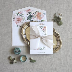 以下が含まれることがあります： 花柄のデザインと繊細なリボンが付いた白い結婚式の招待状。招待状は薄い灰色の背景に、金のトレイと2つの小さなターコイズブルーのリングボックスが置かれています。