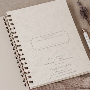 May include: A white journal with a floral design and the text "This journey of happiness & growth belongs to" and "Lovingly handcrafted by Jen and Luke, San Clemente, California &copy;2020 Jen Simpson Design, LLC".