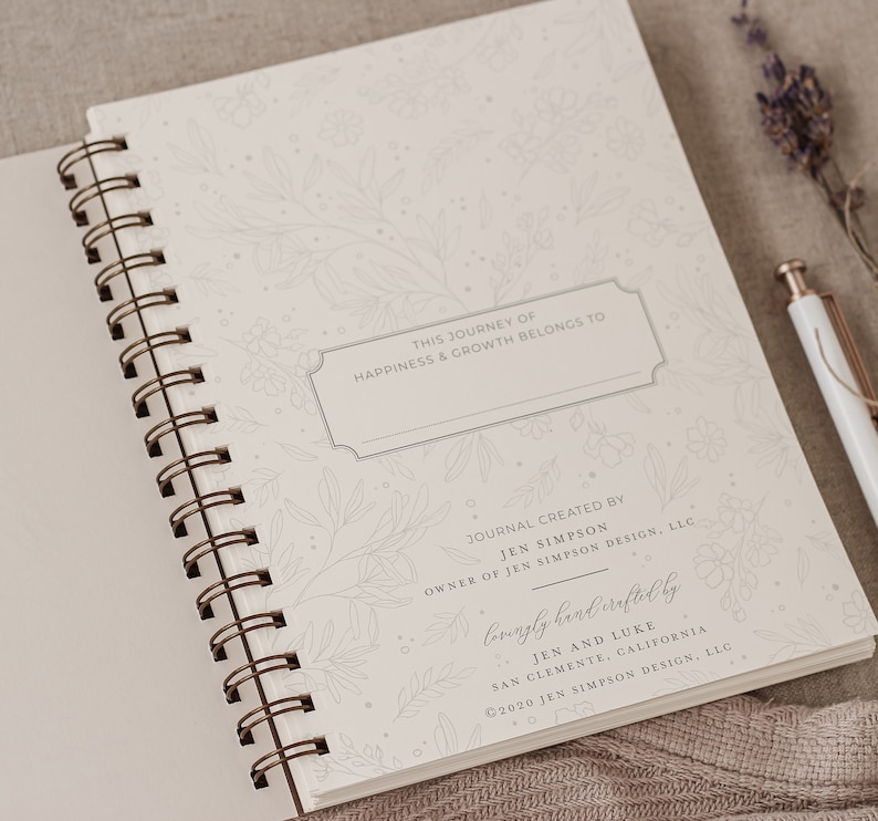 May include: A white journal with a floral design and the text "This journey of happiness & growth belongs to" and "Lovingly handcrafted by Jen and Luke, San Clemente, California &copy;2020 Jen Simpson Design, LLC".
