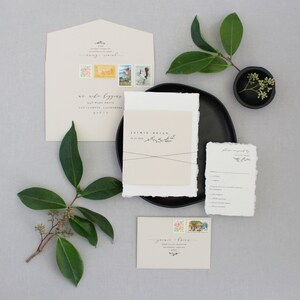 May include: Wedding invitation suite with a simple, elegant design. The invitation features a beige card with the couple's names and wedding date, a small sprig of greenery, and a black ribbon. The RSVP card is a white card with a simple design and the text "Please respond by".
