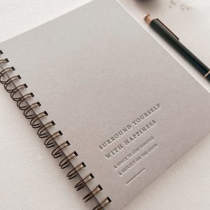 May include: A gray spiral bound journal with the text "Surround Yourself With Happiness" and the subtext "A space to stay positive & reflect on the good".