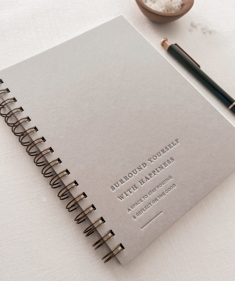 May include: A gray spiral bound journal with the text "Surround Yourself With Happiness" and the subtext "A space to stay positive & reflect on the good".