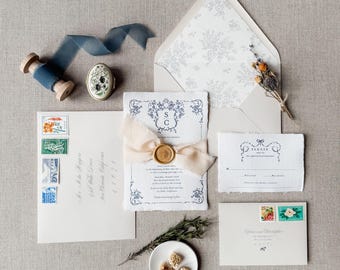 May include: A wedding invitation suite with a blue and white floral design. The invitation features a wax seal with the initials "S.C." and a delicate ribbon. The RSVP card is a separate piece of paper with a matching design. The envelope is lined with a floral pattern.