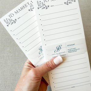 Könnte beinhalten: Zwei weiße 10x23 cm Notizblöcke mit dem Text "TO MARKET" und floralen Akzenten. Jeder Notizblock hat Linien zum Schreiben und einen Abschnitt für Notizen. Die Notizblöcke werden von einem transparenten Band mit dem Text "JEN SIMPSON DESIGN" zusammengehalten.