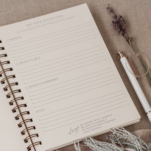 May include: A white spiral bound journal with a cream colored page open to a section titled "Set Your Intentions". The page has lines for writing and includes the text "Let this page guide you through your journey". The journal is on a beige surface with a pen and dried lavender.
