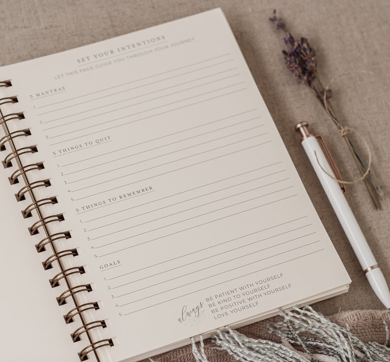 May include: A white spiral bound journal with a cream colored page open to a section titled "Set Your Intentions". The page has lines for writing and includes the text "Let this page guide you through your journey". The journal is on a beige surface with a pen and dried lavender.