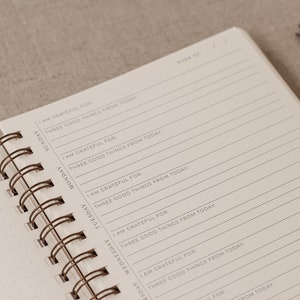 May include: A white spiral bound journal with a brown wire binding. The cover has the text "Week Of" at the top and the days of the week listed down the left side. Each day has the text "I am grateful for: Three good things from today."