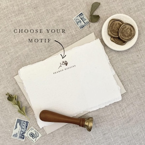 May include: A white rectangular card with deckled edges featuring the printed text SHARON HIGGINS and a small floral icon. The card rests on a light envelope, accompanied by a wooden wax seal stamp, three postage stamps, and wax seals.
